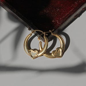 Puede incluir: Un par de pendientes de aro dorados, cada uno con un dise&ntilde;o en forma de coraz&oacute;n. Los pendientes se muestran sobre un fondo gris neutro, con una superficie burdeos oscuro en la parte superior de la imagen.