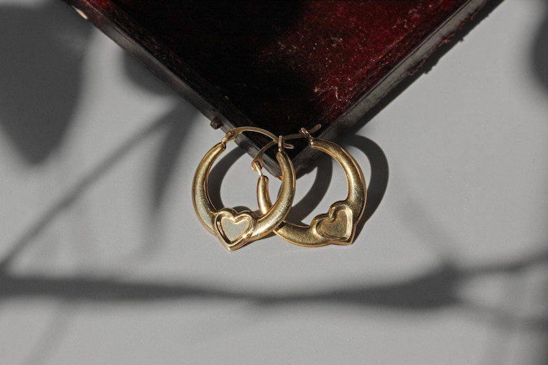 Puede incluir: Un par de pendientes de aro dorados, cada uno con un dise&ntilde;o en forma de coraz&oacute;n. Los pendientes se muestran sobre un fondo gris neutro, con una superficie burdeos oscuro en la parte superior de la imagen.