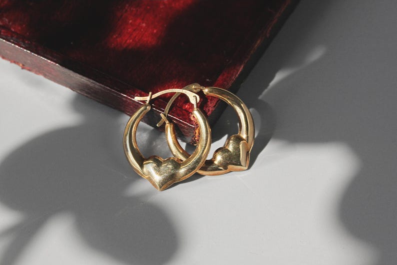 Puede incluir: Un par de pendientes de aro dorados con dijes en forma de coraz&oacute;n. Los pendientes se exhiben sobre una superficie gris claro con un fondo de madera rojo oscuro. Los pendientes est&aacute;n hechos de un material dorado brillante.