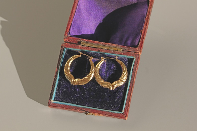 May include: Gold hoop earrings with a textured wave pattern, presented in an open, antique red jewellery box with a purple velvet lining. The earrings are a warm gold colour.