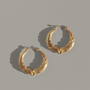 Op de afbeelding: Een paar goudkleurige oorringen met een getextureerd, gevlochten ontwerp. De oorbellen worden gepresenteerd op een grijze ondergrond en werpen schaduwen. De sieraden hebben een klassieke, elegante stijl.
