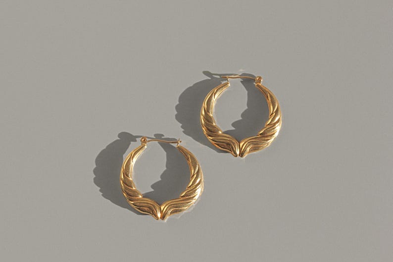 Op de afbeelding: Een paar goudkleurige oorringen met een getextureerd, gevlochten ontwerp. De oorbellen worden gepresenteerd op een grijze ondergrond en werpen schaduwen. De sieraden hebben een klassieke, elegante stijl.