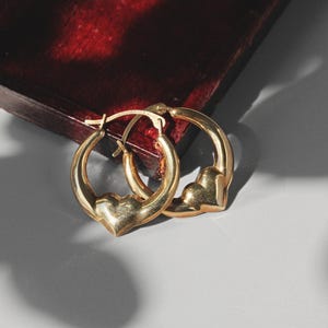 Puede incluir: Un par de pendientes de aro dorados con dijes en forma de coraz&oacute;n. Los pendientes se exhiben sobre una superficie gris claro, con una caja de madera roja oscura en el fondo. Los pendientes son de metal brillante.