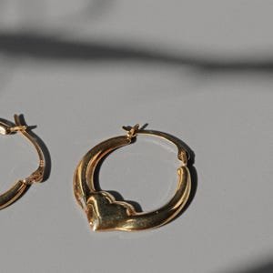 Puede incluir: Dos pendientes de aro dorados con un dise&ntilde;o &uacute;nico en forma de coraz&oacute;n. Los pendientes est&aacute;n hechos de un material met&aacute;lico brillante y se exhiben sobre una superficie gris. Los pendientes son un art&iacute;culo de joyer&iacute;a cl&aacute;sico.