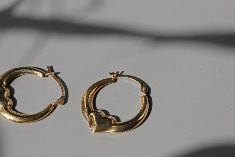 Puede incluir: Dos pendientes de aro dorados con un dise&ntilde;o &uacute;nico en forma de coraz&oacute;n. Los pendientes est&aacute;n hechos de un material met&aacute;lico brillante y se exhiben sobre una superficie gris. Los pendientes son un art&iacute;culo de joyer&iacute;a cl&aacute;sico.