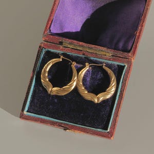 Op de afbeelding: Een paar gouden oorringen met een getextureerd, golfachtig ontwerp, tentoongesteld in een open, antieke sieradendoos. De doos is gevoerd met paars fluweel en heeft een rode en blauwe buitenkant. De oorbellen zijn goudkleurig.