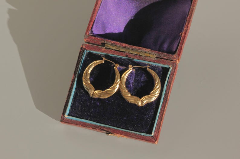 Op de afbeelding: Een paar gouden oorringen met een getextureerd, golfachtig ontwerp, tentoongesteld in een open, antieke sieradendoos. De doos is gevoerd met paars fluweel en heeft een rode en blauwe buitenkant. De oorbellen zijn goudkleurig.