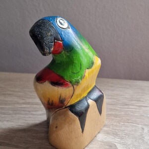 May include: A wooden parrot figurine painted in bright colors, including blue, green, yellow, red, and black. The parrot is perched on a wooden base.
