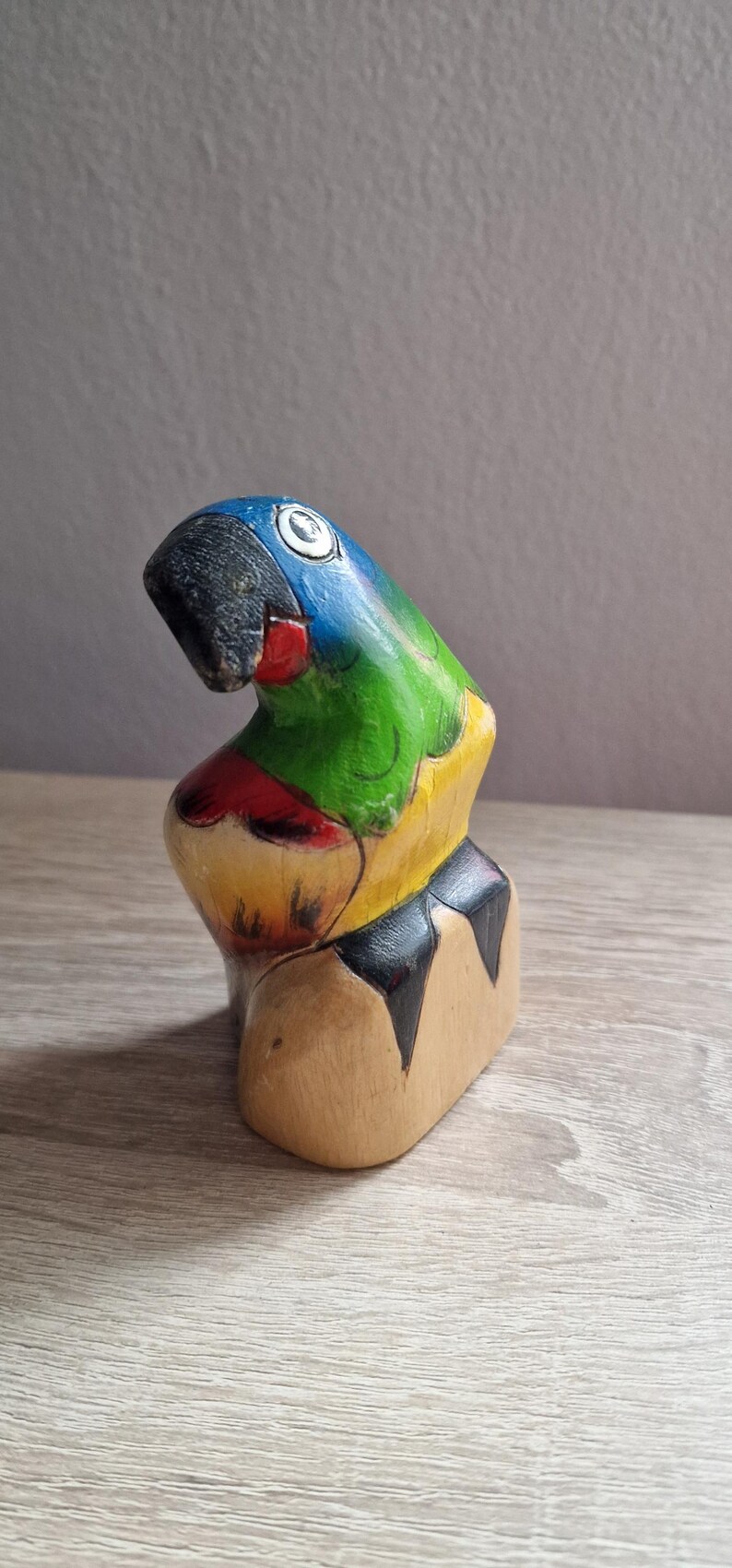 May include: A wooden parrot figurine painted in bright colors, including blue, green, yellow, red, and black. The parrot is perched on a wooden base.