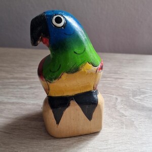 May include: Hand-painted wooden parrot figurine with blue, green, yellow, and black colors. The parrot is perched on a wooden base.