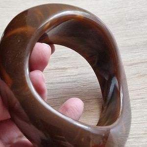 Può includere: Un braccialetto in plastica marrone con una finitura lucida. Il braccialetto ha una forma rotonda e liscia e un design semplice.