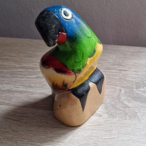 May include: Hand-painted wooden parrot figurine with blue, green, yellow, red, and black colors. The parrot is perched on a wooden base.