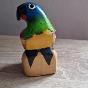 May include: A wooden parrot figurine with blue, green, yellow, and black paint. The parrot is perched on a wooden base.