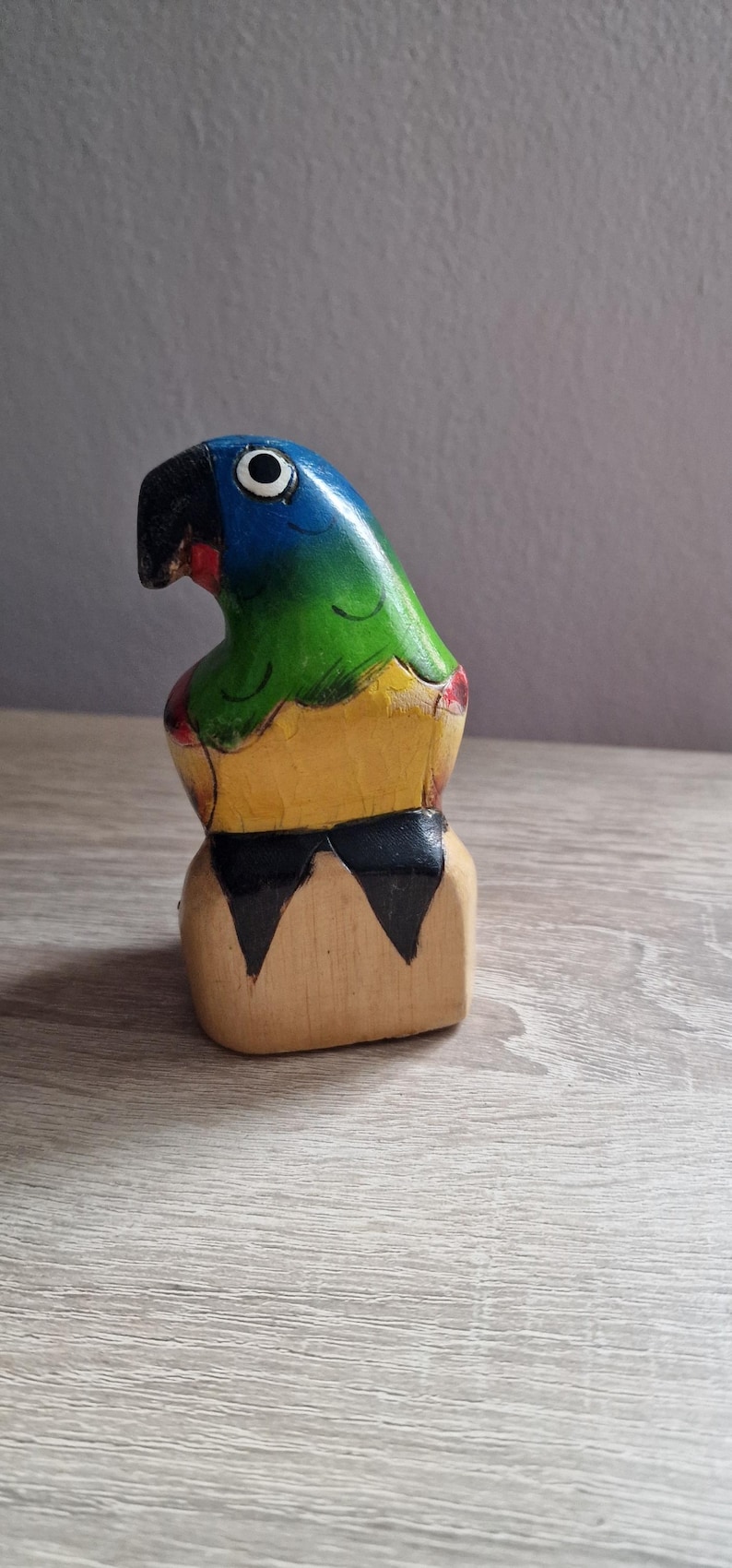 May include: A wooden parrot figurine with blue, green, yellow, and black paint. The parrot is perched on a wooden base.