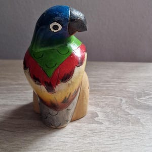 May include: A wooden parrot figurine painted in blue, green, red, and yellow. The parrot has a black beak and a white eye.