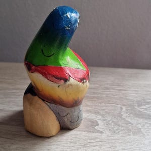 May include: A colorful wooden figurine with a blue, green, red, yellow, and gray paint scheme. The figurine is shaped like a person with a rounded head and a body that is bent over.