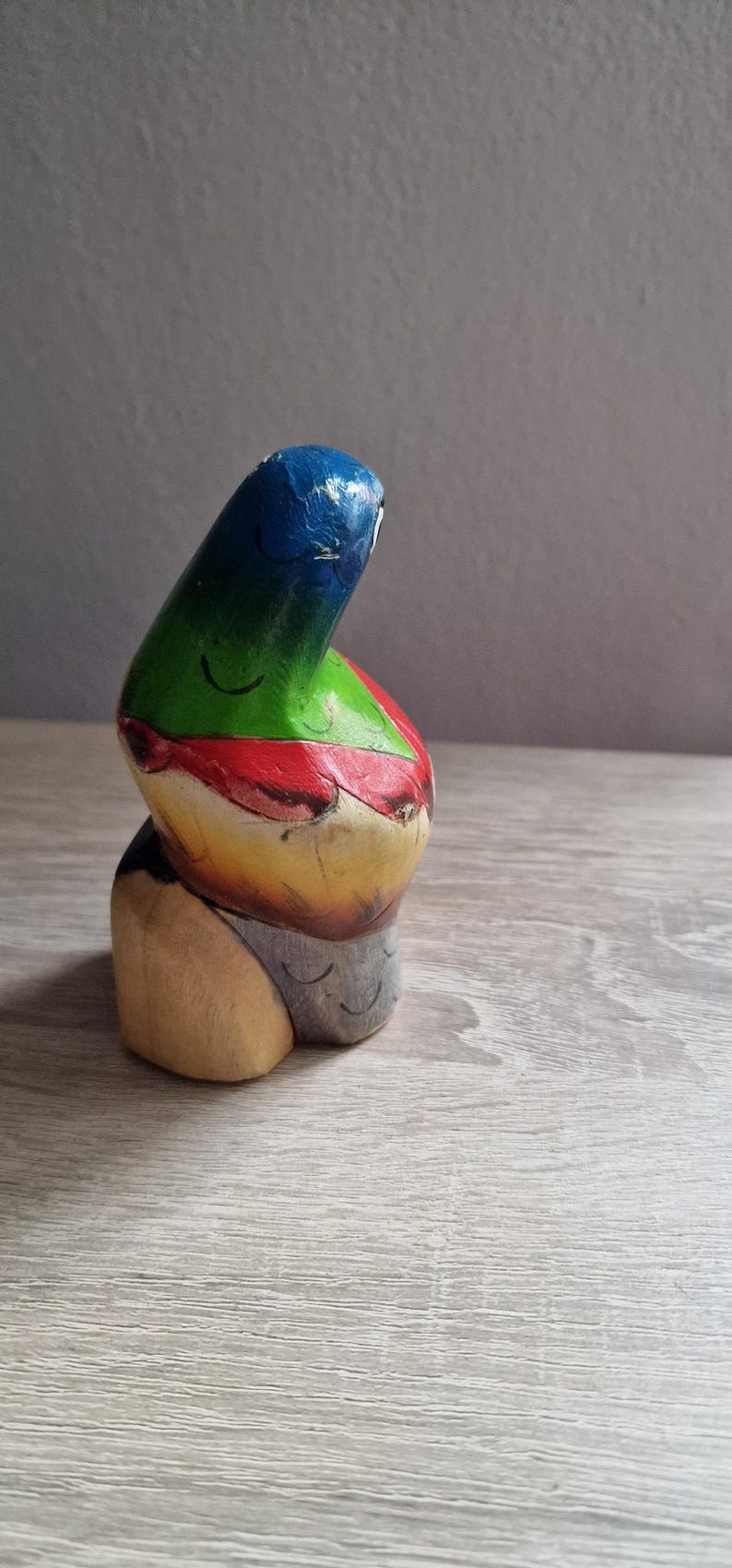 May include: A colorful wooden figurine with a blue, green, red, yellow, and gray paint scheme. The figurine is shaped like a person with a rounded head and a body that is bent over.