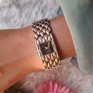 Puede incluir: Un reloj de pulsera plateado con una correa de eslabones texturizados en forma de escamas y una esfera cuadrada negra. La esfera muestra el texto "ESQ" y "SWISS" en blanco, con manecillas plateadas que indican la hora sobre un fondo oscuro.