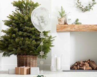 May include: A Christmas scene featuring a decorated evergreen tree in a wicker basket, a clear balloon with snowflake designs, and a string of snowflake ornaments. Several wrapped gifts and a candle in a glass holder are also present.