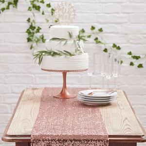 Op de afbeelding: Een drielaagse witte bruidstaart met een houten "Mr & Mrs"-topper staat op een gouden taartstandaard. De taart is versierd met groen en staat op een tafel met een roze gouden sequintafelkleed.