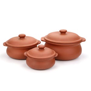 Puede incluir: Un juego de tres ollas de terracota con tapas a juego. Cada olla presenta un cuerpo redondeado, dos pequeñas asas laterales y un pomo central en la tapa. El material de arcilla tiene un acabado natural de color marrón rojizo mate.