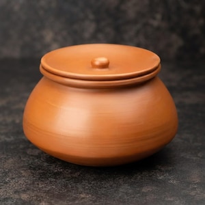 Puede incluir: Una vasija de barro terracota con un cuerpo liso y redondeado y una tapa circular a juego. La tapa presenta un pequeño pomo central como asa. La vasija tiene una forma ancha y bulbosa con un acabado mate en tonos tierra naturales.