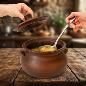 May include: A round, dark brown clay pot with two small side handles and a matching lid. The pot contains a light-colored soup or stew, and a long-handled silver metal ladle is positioned inside the pot to scoop the liquid contents.
