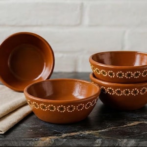 May include: A set of four terracotta bowls with a smooth, glossy finish. Each bowl features a decorative band of small, white, flower-like patterns encircling the exterior. The bowls are arranged in a cluster, with one standing upright and three stacked.