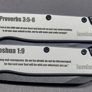 Könnte beinhalten: Zwei silberne Klappmesser mit schwarzen Akzenten. Jedes Messer trägt einen Bibelvers und den Markennamen "Kershaw". Auf einem Messer steht "Sprüche 3:5-6" und auf dem anderen "Josua 1:9".