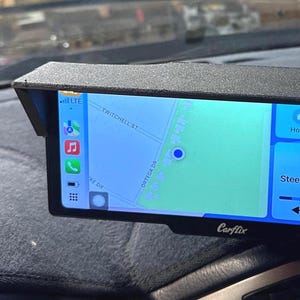 May include: A black Carflix device displaying a map and music controls. The screen shows a map with street names and a music player with the song "Steel Rail Blues" by Gordon Lightfoot. The device is mounted on a car dashboard.