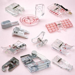 May include: A collection of clear plastic sewing machine presser feet, including various styles for different stitching techniques. Some feet have red markings or grid patterns. The items are arranged on a light pink surface.