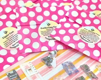 May include: A collection of metal and plastic sewing machine presser feet, including a zipper foot, gathering foot, and bias binding foot, arranged in front of pink and white polka dot paper envelopes with printed instructional labels.