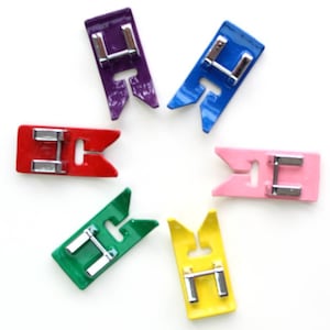 May include: Six colourful sewing machine presser feet with metal bars. The feet are in a starburst pattern and are red, green, yellow, pink, blue, and purple.
