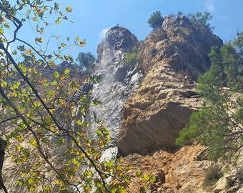 May include: A rocky cliff face with a waterfall cascading down the side. Lush green trees frame the scene, with some leaves turning yellow.