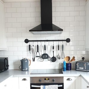 May include: A modern kitchen with white cabinets, a black range hood, and a white tiled backsplash. Kitchen utensils hang above the stove. A gray toaster and kettle sit on the countertop. The floor is light brown.