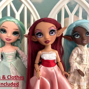 May include: Three fantasy dolls with pointed ears and detailed makeup. The dolls have different skin tones and hair colors: mint green, burgundy, and light blue. They are dressed in elegant gowns with lace and satin details. The text at the bottom reads "Dolls & Clothes Not Included."