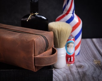 Puede incluir: Una bolsa de tocador de cuero marrón con cierre de cremallera y asa de cuero. La bolsa está sobre una superficie negra junto a un cepillo de afeitar blanco, una botella de aftershave y una lata de crema de afeitar.