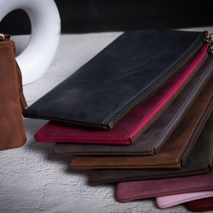 May include: A stack of nine leather zippered pouches in various colors, including brown, black, burgundy, gray, and pink. The pouches are arranged in a staggered pattern, with the top pouch being brown and the bottom pouch being pink.