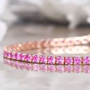 Puede incluir: Una pulsera de oro rosa con una fila de gemas rosas vibrantes. Las gemas están engastadas muy juntas, creando una línea continua de color. La pulsera se muestra sobre una superficie reflectante.
