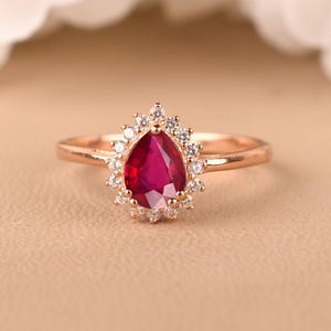 May include: A rose gold ring featuring a pear-shaped deep red gemstone at the centre, surrounded by a halo of small, clear, round-cut stones. The band is thin and polished, showcasing a classic design with a vibrant, saturated red focal point.