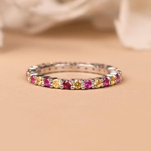 May include: A silver eternity ring featuring alternating pink and yellow gemstones. The ring is set against a soft, neutral background, with a blurred white floral element in the background.