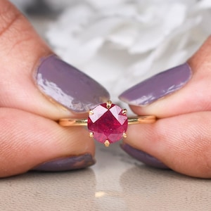 Puede incluir: Un anillo de oro con una gran gema facetada de color rojo intenso. El anillo tiene una fina banda de oro pulido. La gema está sujeta por seis delicadas garras. El anillo se sostiene entre dos manos con esmalte de uñas morado.