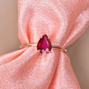 May include: A teardrop-shaped ruby ring set in a gold band. The ring is on a pink satin fabric.