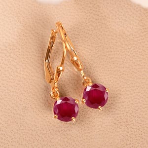 May include: A pair of gold earrings with red ruby gemstones. The earrings have a simple design with a small, round ruby dangling from a gold hoop.