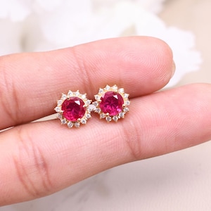 May include: A pair of gold stud earrings with a red gemstone surrounded by smaller white gemstones.
