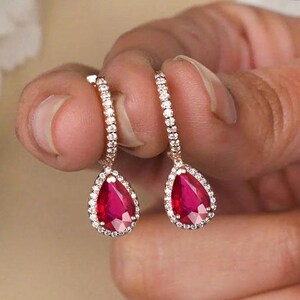 Peut inclure: Une paire de boucles d'oreilles pendantes en or rose avec des pierres précieuses rouges en forme de poire. Chaque boucle est ornée d'un halo de petites pierres blanches autour de la gemme centrale et d'une rangée de pierres sur l'attache.