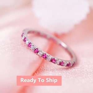 May include: A thin silver-toned band ring featuring an alternating pattern of small, round, brilliant-cut pink rubies and clear white diamonds set along the entire circumference of the metal band. The text Ready To Ship is visible below.