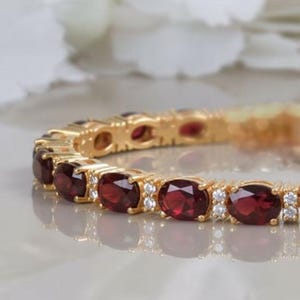 Könnte beinhalten: Ein goldfarbenes Armband mit einer Reihe von oval geschliffenen, tiefroten Granat-Edelsteinen, die sich mit kleinen, klaren, rund geschliffenen weißen Steinen abwechseln. Die Metallfassung ist poliert und gelbgoldfarben.