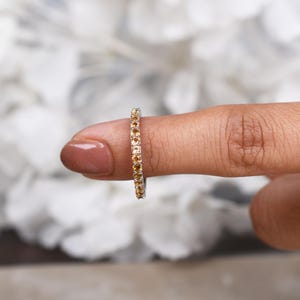 May include: A gold ring set with a continuous line of small, round, amber-coloured gemstones. The ring is on a finger, with a blurred white floral background.