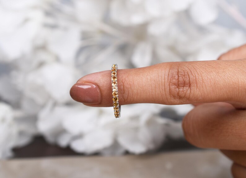 May include: A gold ring set with a continuous line of small, round, amber-coloured gemstones. The ring is on a finger, with a blurred white floral background.